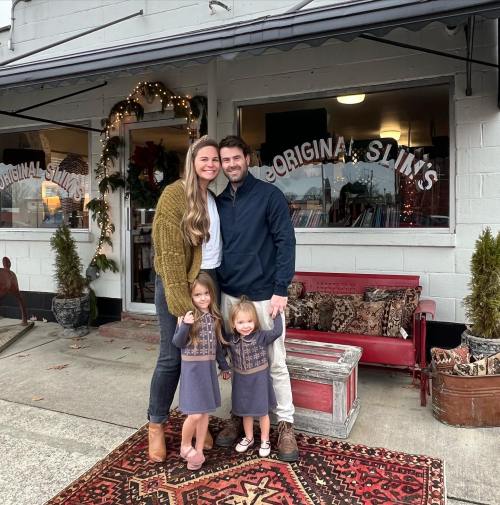 A family of four stands outside a shop, smiling. Two young girls in matching dresses pose in front of their parents.