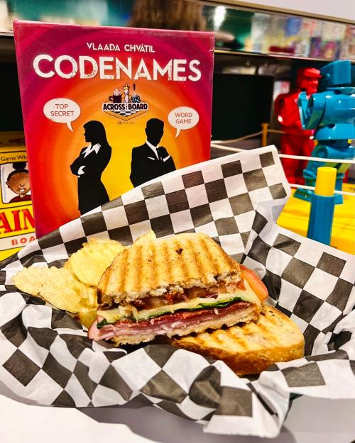 A grilled sandwich with chips in a checkered basket, next to the board game "Codenames" on a colorful table.
