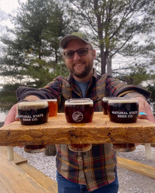A person holds a wooden flight board with several beer samples, smiling in front of trees and a cloudy sky.