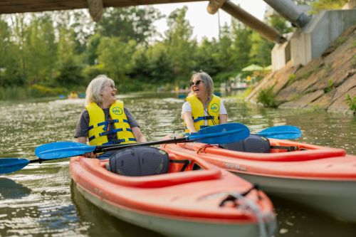 Two women in kayaks wearing life jackets smile at each other on a calm river surrounded by greenery.