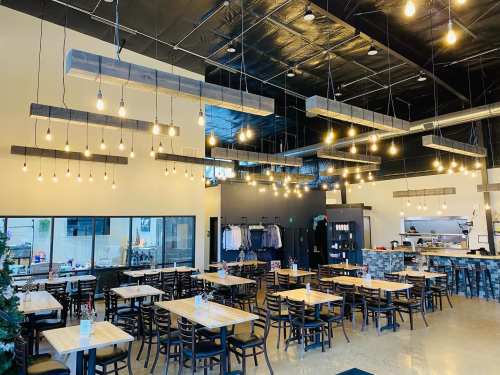 A modern restaurant interior with wooden tables, black chairs, and hanging light fixtures, featuring an open kitchen area.