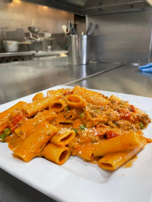 A plate of rigatoni pasta with a creamy sauce, cherry tomatoes, peas, and grated cheese, set in a kitchen environment.