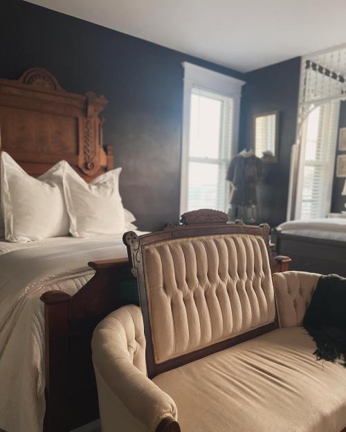 Cozy bedroom featuring a vintage bed, a tufted loveseat, and natural light from windows. Dark walls create a warm atmosphere.