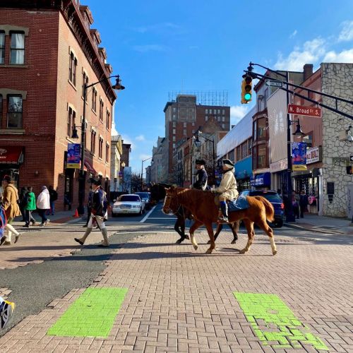 Two people on horseback cross a city street, with pedestrians and buildings in the background under a clear blue sky.