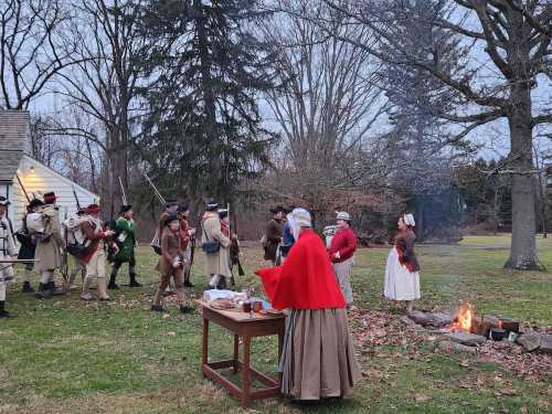 Historical reenactors in period costumes gather around a fire, preparing food and engaging in activities outdoors.