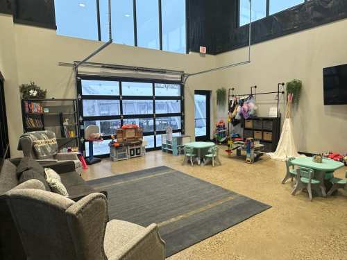 A spacious playroom with toys, small tables, a cozy seating area, and large windows letting in natural light.