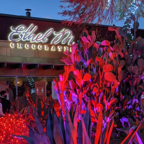 Brightly lit storefront of Ethel M Chocolates, surrounded by colorful plants and festive decorations at dusk.