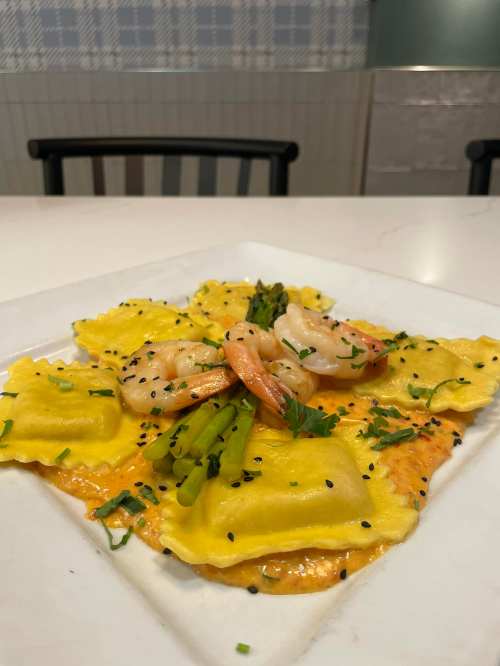 Plate of yellow ravioli topped with shrimp, asparagus, and black sesame seeds, garnished with chopped herbs.