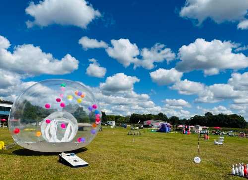 A large transparent inflatable bubble with colorful balls inside, set in a grassy field under a blue sky with fluffy clouds.