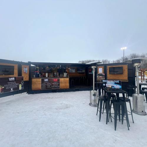 A winter outdoor bar with wooden accents, tables, and heaters, set on a snowy landscape under a gray sky.