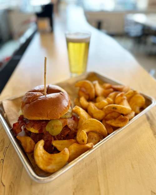 A crispy chicken sandwich with toppings, served with golden potato wedges and a glass of beer on a wooden table.