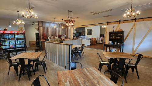 A cozy café interior with wooden tables, metal chairs, a counter, and shelves filled with drinks and snacks.