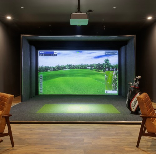 Indoor golf simulator with a large screen displaying a green course, golf clubs nearby, and seating in the foreground.