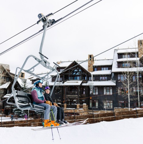 Two skiers on a ski lift, surrounded by snow and a mountain lodge in the background.