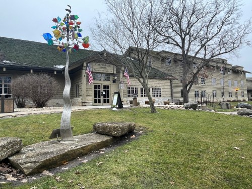 A building with a stone facade, colorful sculpture, and trees, set in a grassy area with rocks and flags.