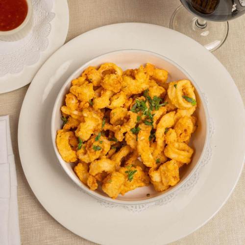 A bowl of golden, crispy shrimp garnished with parsley, served on a white plate with a glass of wine in the background.