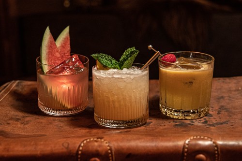 Three colorful cocktails on a wooden surface: a watermelon drink, a mint garnish cocktail, and a golden drink with a raspberry.