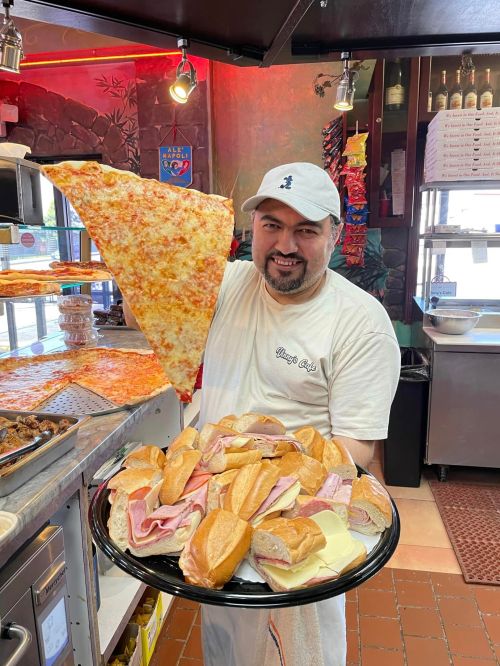 A smiling man holds a giant slice of pizza in one hand and a platter of assorted sandwiches in the other.