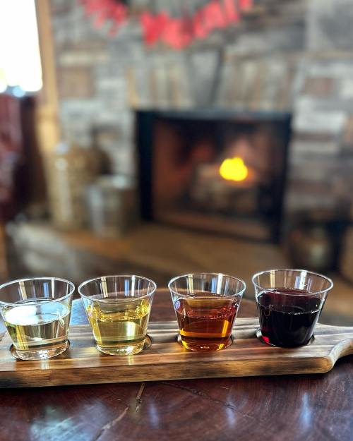 Four glasses of different colored beverages on a wooden tray, with a cozy fireplace in the background.