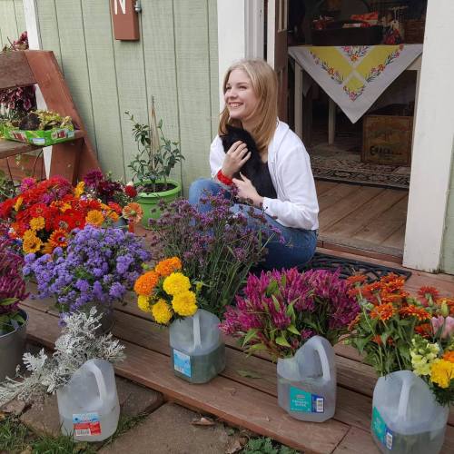 A smiling woman sits on a porch surrounded by colorful flowers and holds a black cat.