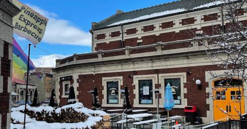 A snowy outdoor dining area with tables, a barbecue sign, and colorful flags near a brick building.