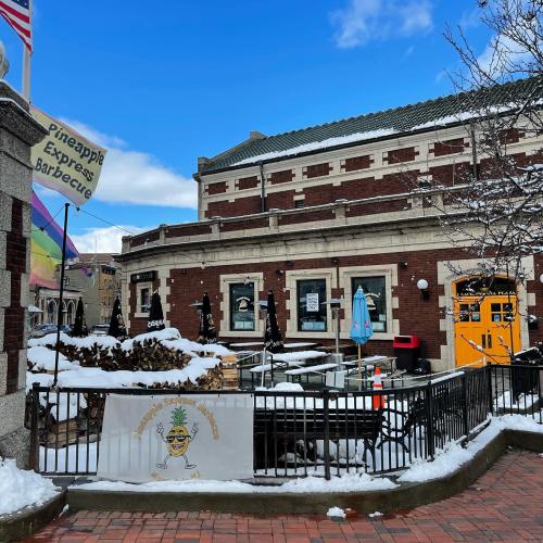 A snowy street scene featuring a barbecue restaurant with outdoor seating and colorful signage.