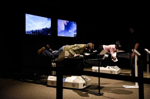Two people wearing VR headsets lie on devices, interacting with screens displaying landscapes, in a dark room.