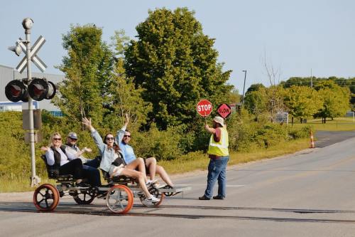 A group of four people on a cart wave while a man in a vest holds a stop sign at a railroad crossing.