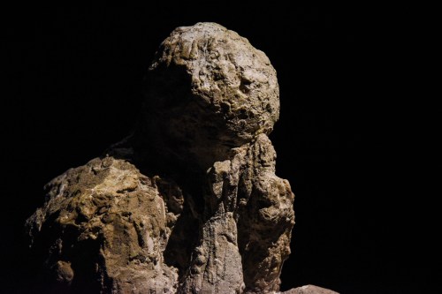A textured stone sculpture of a figure sitting with hands on its face, set against a dark background.