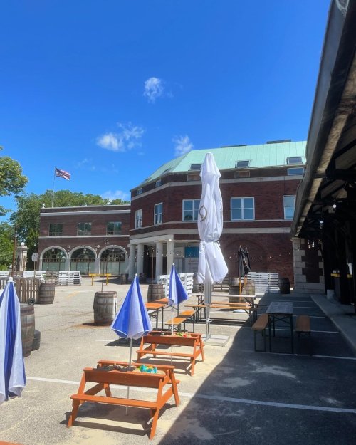 Outdoor seating area with umbrellas in front of a brick building under a clear blue sky. American flag visible.