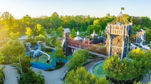 Aerial view of a vibrant playground with slides, towers, and lush greenery under a bright, sunny sky.