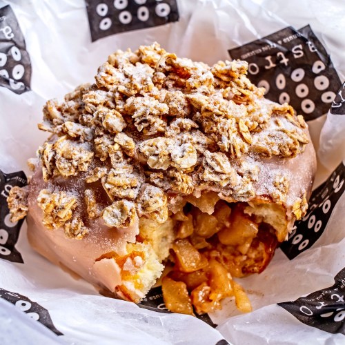A glazed donut topped with crunchy granola and filled with apple filling, resting on patterned paper.