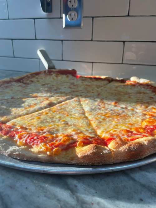 A freshly baked cheese pizza on a metal tray, with slices cut and sunlight casting shadows on the surface.
