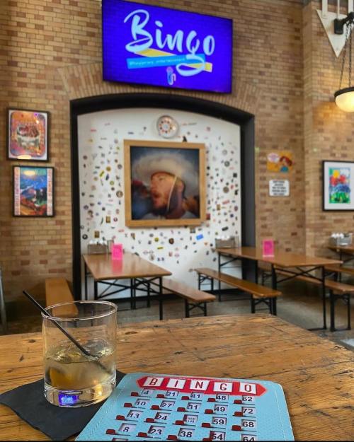 A bingo card and drink sit on a table in a lively venue with a "Bingo" sign and colorful decor in the background.