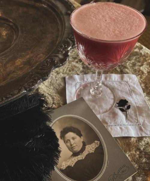 A vintage photo of a woman beside a pink cocktail in an elegant glass on a textured surface.