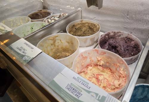 A display of various ice cream flavors in tubs, including strawberry cheesecake and others, in a shop setting.