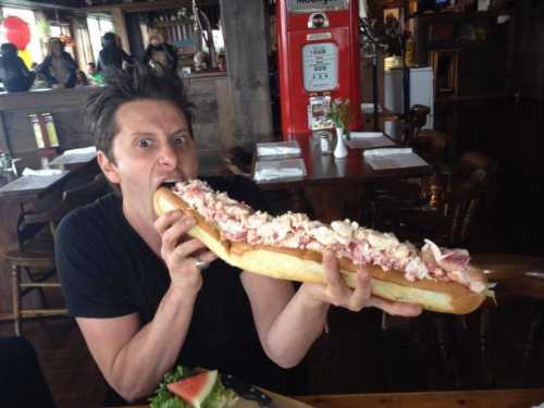 A person excitedly prepares to take a big bite of an oversized hot dog topped with creamy ingredients.