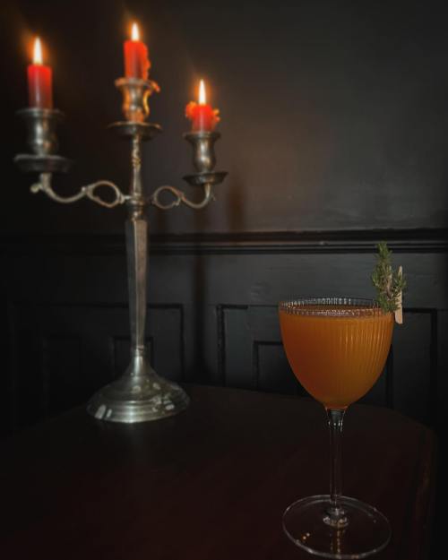 A cocktail in a vintage glass sits on a table beside a silver candelabra with lit red candles against a dark background.