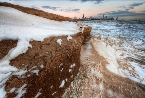 A sandy shoreline with snow and ice, overlooking a city skyline under a cloudy sky at dusk.