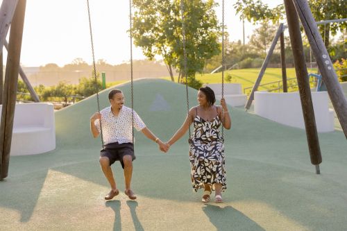 A couple sits on swings in a park, holding hands and smiling, with greenery and sunlight in the background.