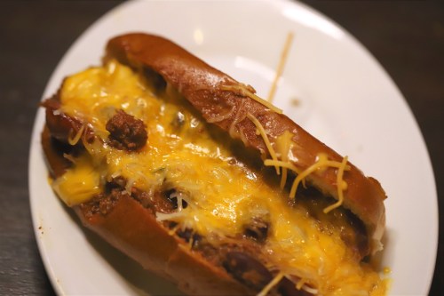 A cheesy chili hot dog topped with shredded cheese on a white plate.
