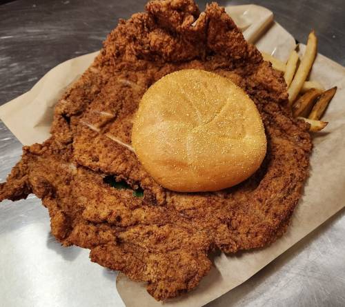 A large, crispy fried chicken breast served with a bun and fries on a paper-lined plate.