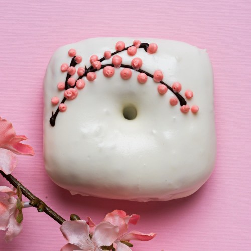 A white donut decorated with pink floral icing, placed on a pink background with cherry blossom petals nearby.