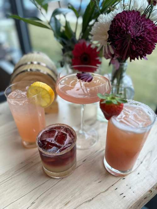 Four colorful cocktails on a wooden table, surrounded by fresh flowers in vases.