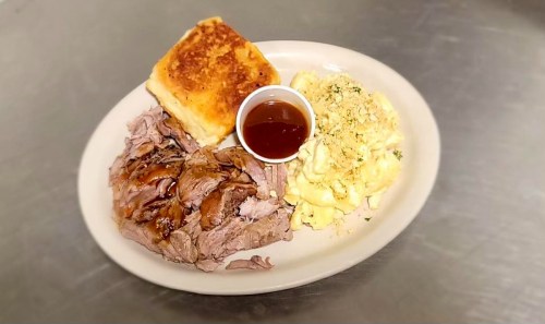 A plate of sliced barbecue meat, a biscuit, creamy macaroni and cheese, and a small cup of sauce.