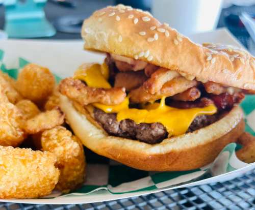 A close-up of a cheeseburger with crispy onion rings and bacon, served with a side of golden tater tots.