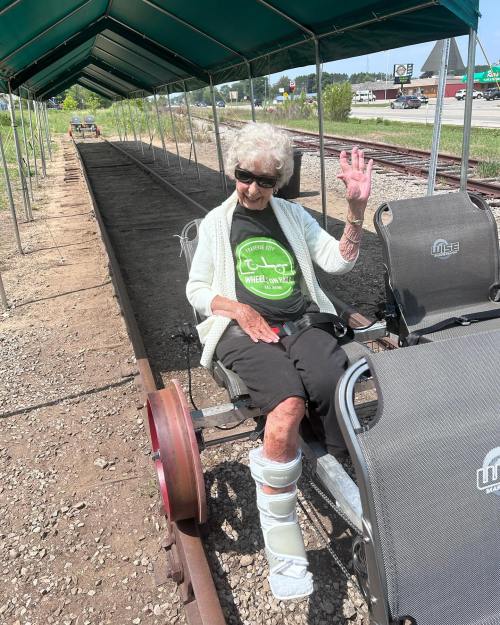 An elderly woman in a black t-shirt sits on a rail cart, smiling and waving, under a green canopy.