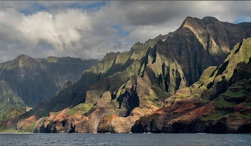 Lush green mountains rise dramatically from the ocean under a cloudy sky, showcasing rugged cliffs and vibrant colors.
