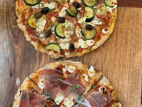Two pizzas on a wooden surface: one topped with zucchini, olives, and cheese; the other with prosciutto and herbs.