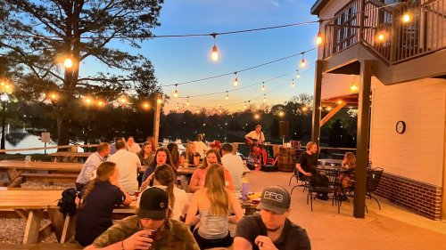 A lively outdoor dining scene at sunset, with string lights, people enjoying food, and a musician performing.